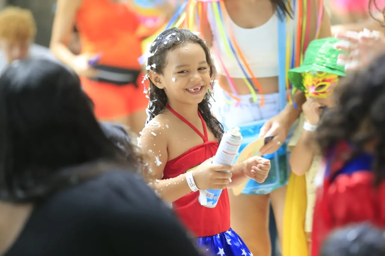 Menina de blusa vermelha e saia azul com serpentina no cabelo e segurando espuminha.