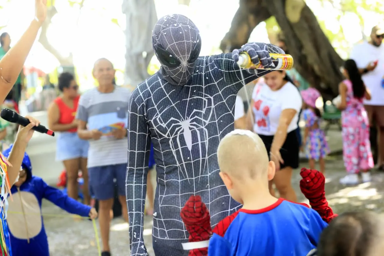 Pessoa fantasiada de Homem Aranha com traje preto, espirrando espuminha em uma criança.