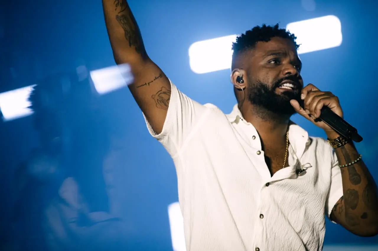 Cantor em momento de performance, com braço levantado e expressão intensa, sob iluminação azul e branca de palco.