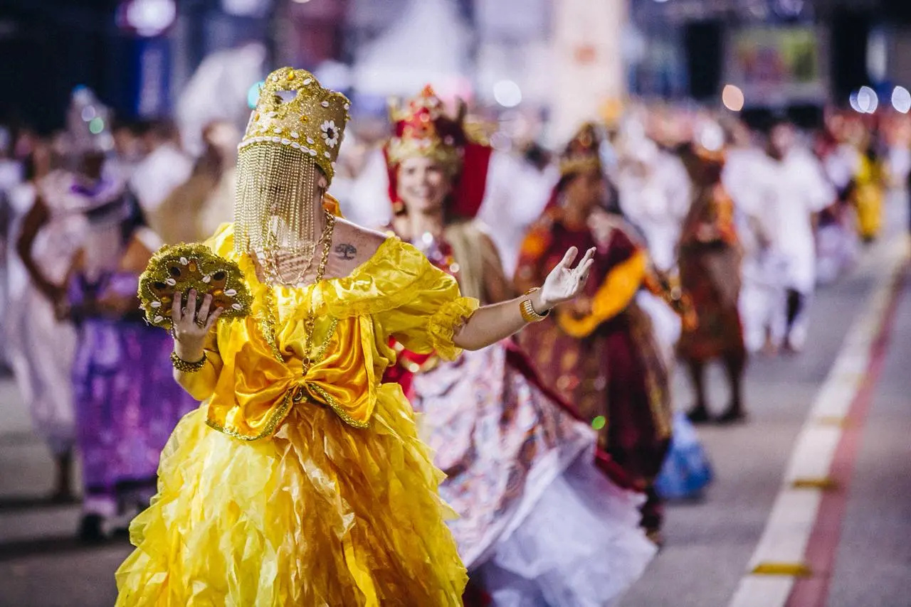 Integrante de Maracatu desfila à noite com traje amarelo volumoso e brilhante, usando coroa ornamentada com véu de miçangas que cobre o rosto e segurando leque decorado; ao fundo, outros participantes do cortejo seguem pela via pública iluminada.