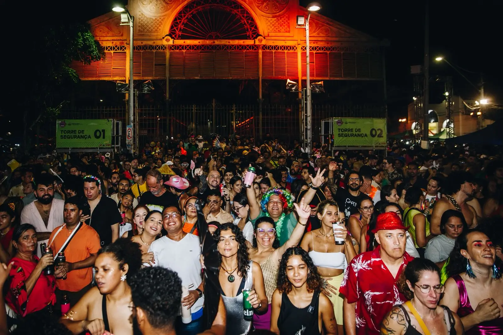 Grande público reunido em frente a prédio histórico iluminado em tons alaranjados; placas indicam “Torre de Segurança 01” e “Torre de Segurança 02”.