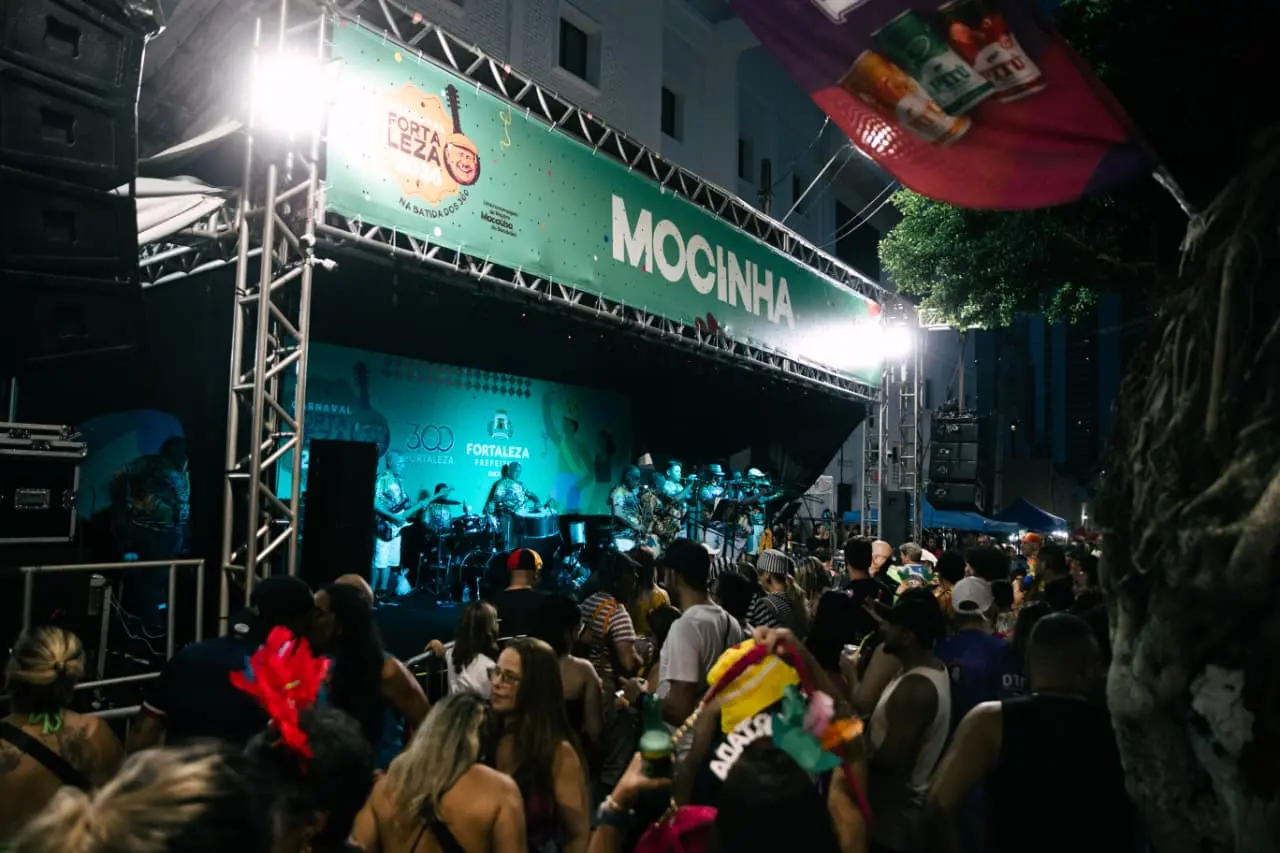 Palco identificado com a palavra “Mocinha” recebe banda durante apresentação noturna; público acompanha o show em frente à estrutura metálica iluminada com banners do Carnaval de Fortaleza.