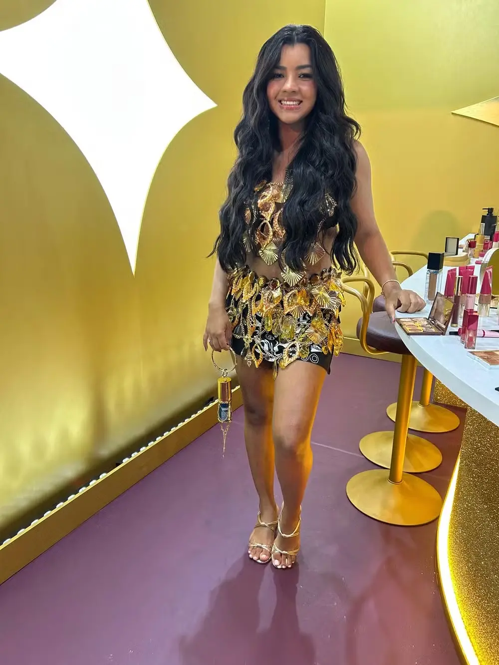 Ary Mirelle, com cabelos pretos longos soltos e roupa brilhosa dourada.