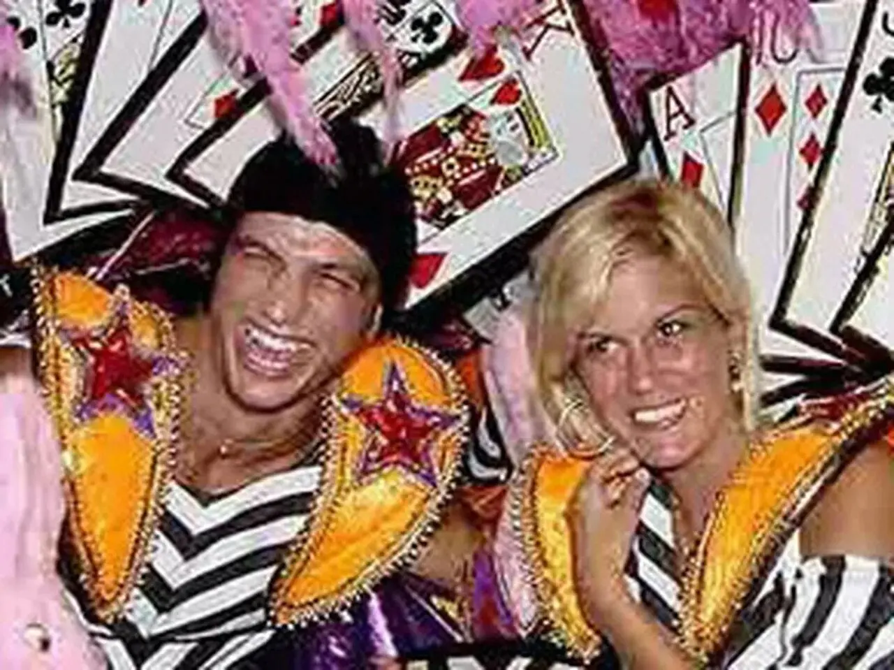 Homem e mulher brancos, ele de bandana preta com adereço rosa e ela de cabelo loiro curto solto, curtindo Carnaval.