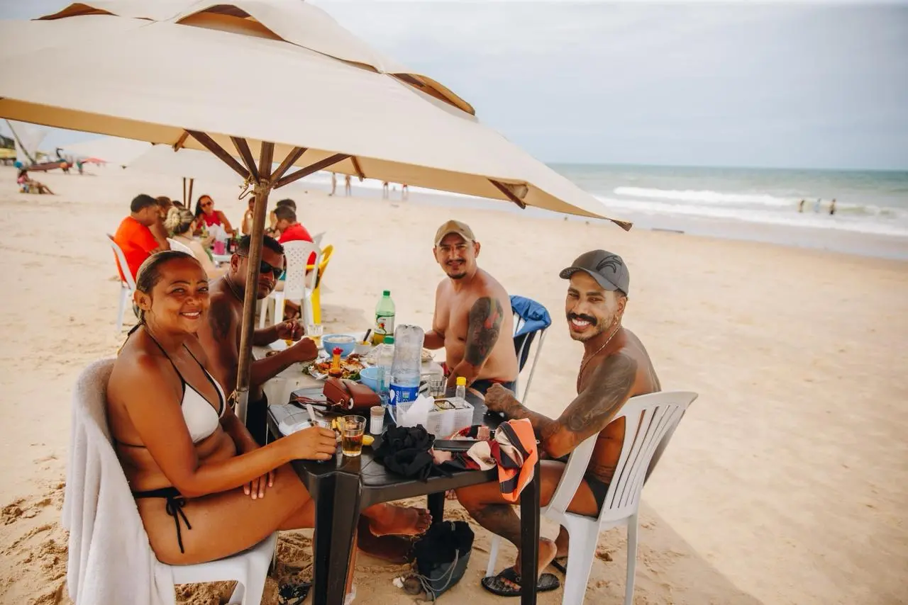 Um grupo de pessoas sentadas em cadeiras de praia ao redor de uma mesa plástica escura sob um grande guarda-sol na areia. Sobre a mesa há garrafas de bebida, copos, embalagens de alimentos e objetos pessoais. O mar aparece ao fundo, com algumas pessoas na água, enquanto outros grupos estão sentados mais afastados na praia. O clima é descontraído e típico de um dia de lazer à beira-mar.