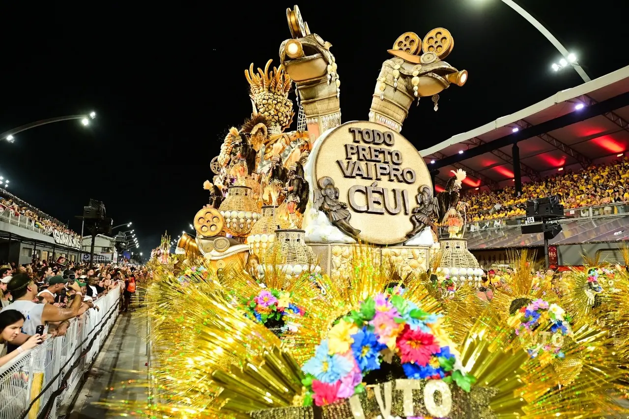 Carro alegórico de escola de samba avança pela avenida durante um desfile noturno, repleto de esculturas douradas, figuras mitológicas e elementos brilhantes. Na parte frontal, há um grande disco com a frase “Todo preto vai pro céu”. À frente do carro, adereços com flores coloridas e franjas douradas ocupam o primeiro plano. Arquibancadas cheias de espectadores aparecem dos dois lados da avenida, iluminadas pelos refletores do sambódromo.