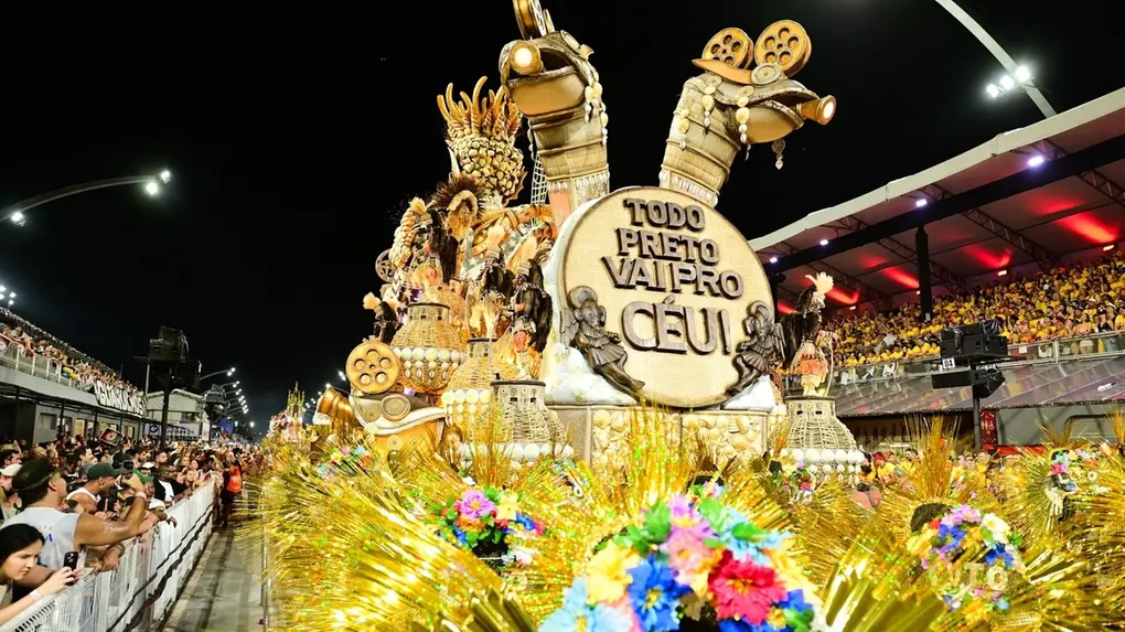 Carro alegórico de escola de samba avança pela avenida durante um desfile noturno, repleto de esculturas douradas, figuras mitológicas e elementos brilhantes. Na parte frontal, há um grande disco com a frase “Todo preto vai pro céu”. À frente do carro, adereços com flores coloridas e franjas douradas ocupam o primeiro plano. Arquibancadas cheias de espectadores aparecem dos dois lados da avenida, iluminadas pelos refletores do sambódromo.
