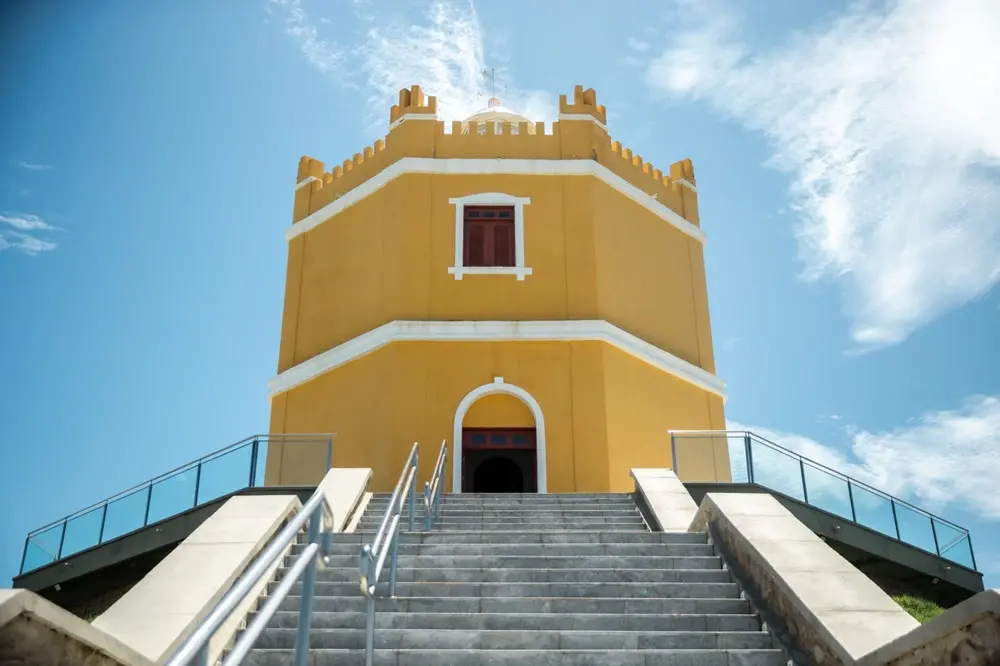 Na imagem, uma fotografia em ângulo baixo (contra-plongée) do Farol do Mucuripe, em Fortaleza. A estrutura octogonal de cor amarela intensa domina o centro da composição, destacando-se contra um céu azul claro com nuvens esparsas. No topo do edifício, veem-se ameias que lembram uma fortificação e a cúpula branca da lanterna com uma biruta metálica. No centro da parte inferior, uma larga escadaria de pedra cinza sobe em direção à entrada principal, que possui um arco branco. Corrimãos metálicos duplos e modernos dividem a escada e margeiam as laterais, que também contam com guarda-corpos de vidro. A perspectiva acentua a imponência do monumento histórico restaurado.