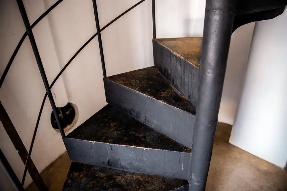 Na imagem, close-up do interior do farol mostrando uma escada caracol de metal preto. Os degraus triangulares apresentam marcas de desgaste e ferrugem dourada, revelando a idade da estrutura. A escada é sustentada por um eixo central cilíndrico preto e possui um corrimão fino de barras metálicas à esquerda. As paredes ao fundo são brancas e simples, criando um contraste industrial com o metal escuro da escadaria.