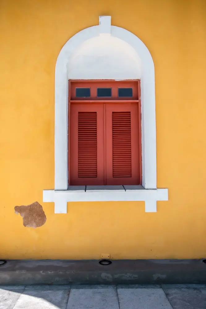 Na imagem, uma janela de madeira em tons de terracota centralizada em uma parede de cor amarelo-mostarda. A janela possui venezianas fechadas na parte inferior e três pequenos painéis retangulares de vidro no topo. Ela é emoldurada por um arco branco em relevo, com detalhes geométricos na base e no topo. Na parte inferior esquerda da parede amarela, há uma pequena área onde o reboco está descascado, revelando a textura marrom por baixo. O chão à frente é composto por placas de pedra cinza.