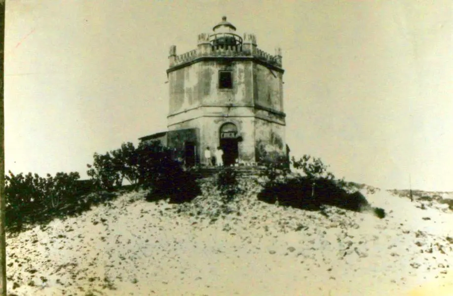 Fotografia antiga em preto e branco do Farol do Mucuripe, mostrando o farol em estrutura octogonal de alvenaria sobre uma duna de areia, cercado por vegetação rasteira e arbustos, sob céu claro.
