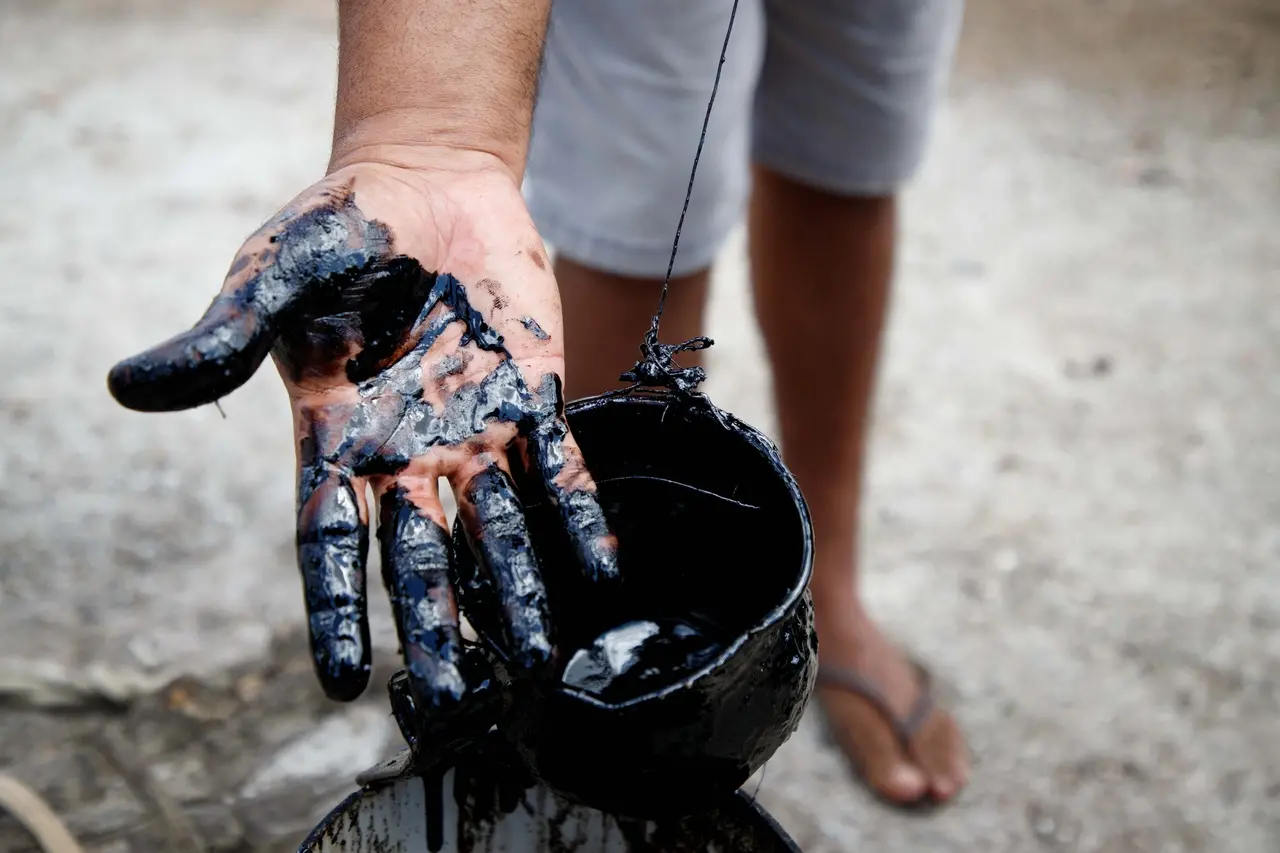 Imagem mostra pessoa segurando uma mão coberta de petróleo, com um balde cheio do material na frente, destaque para a prática de arte com tinta na mão.
