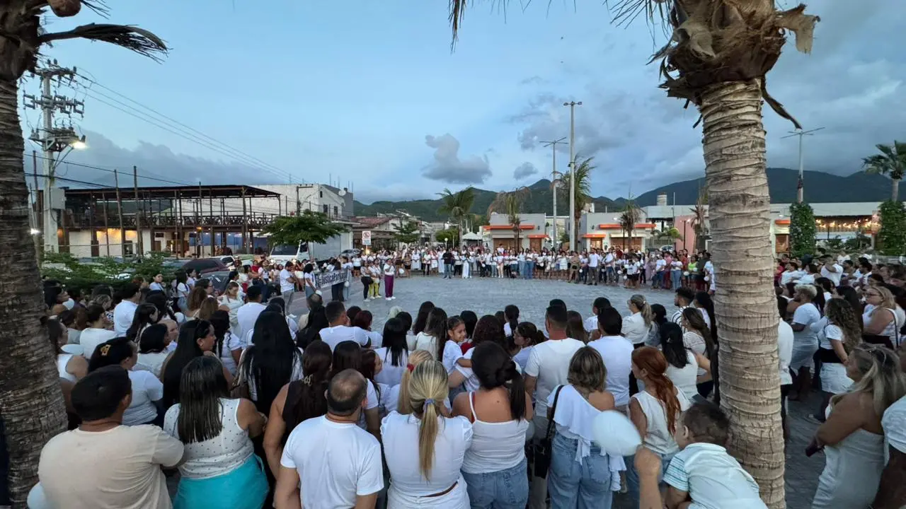 A imagem mostra uma grande concentração de pessoas reunidas em uma praça ao ar livre, formando um círculo em torno de alguém que parece discursar ao centro. A maioria veste roupas claras, predominando o branco — algo comum em atos simbólicos por paz e justiça. O cenário inclui palmeiras, construções ao redor da praça e montanhas ao fundo, sob um céu já no início da noite. O clima aparenta ser de manifestação pacífica, com forte mobilização comunitária.