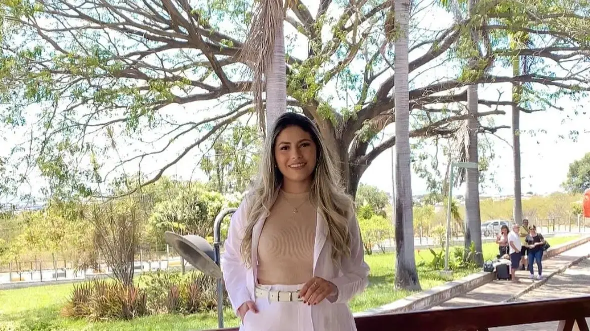 A imagem mostra Ana Karolina, mulher jovem, de cabelos longos e claros, sorrindo enquanto posa ao ar livre. Ela veste calça e blazer brancos, combinados com uma blusa bege. Ao fundo, há árvores de grande porte, palmeiras e uma área verde que parece ser um parque ou praça. Algumas pessoas aparecem mais atrás, próximas a um caminho pavimentado.