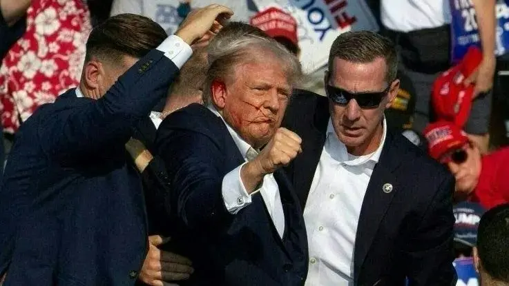 Trump é conduzido por agentes federais após ser alvo de tiro durante a campanha eleitoral. Ele tem manchas de sangue no rosto e posa com os punhos cerrados para cima.