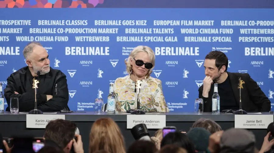 O cineasta Karim Aïnouz, a atriz Pamela Anderson e o ator Jamie Bell participam de uma coletiva de imprensa no Festival de Berlim, sentados atrás de uma bancada com identificações. O trio compõe uma cena profissional e sofisticada sob um fundo azul repleto de logotipos do evento, enquanto interagem diante da audiência.