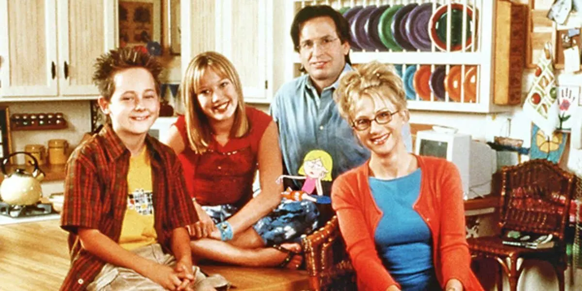 Família reunida na cozinha, sorrindo e ao redor de uma mesa, em um momento de alegria e convivência familiar, com cozinhas ao fundo.