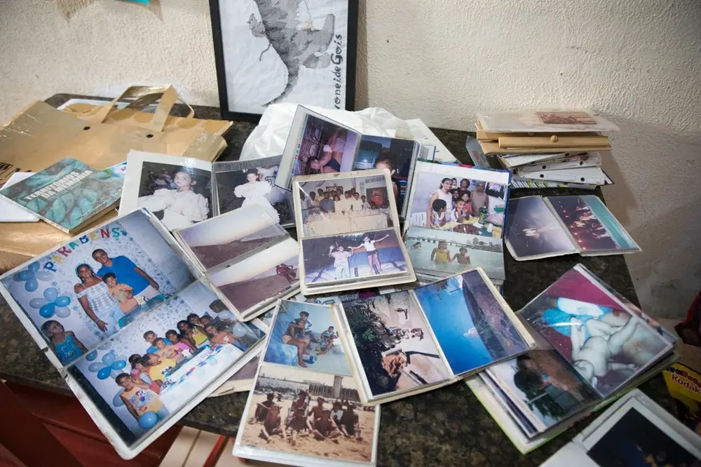 Na imagem, uma mesa de mármore escuro repleta de álbuns de fotografias abertos e fotos avulsas, revelando memórias de uma família. As imagens mostram festas de aniversário com balões, crianças em trajes de gala, momentos na praia e reuniões familiares. No canto superior esquerdo, há uma sacola dourada e um livro intitulado 