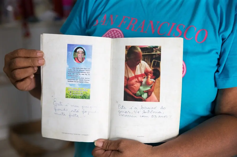Na imagem, uma pessoa segura um pequeno livro aberto com as duas mãos. Nas páginas, há registros de memória de familiares falecidos. A página esquerda contém um 