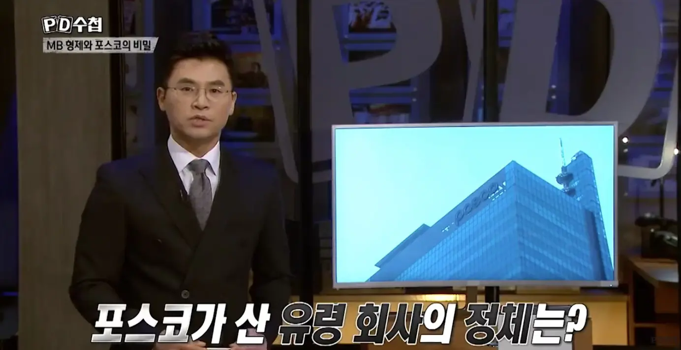 Apresentador de telejornal vestindo terno escuro e gravata, falando diante das câmeras em estúdio. Ao lado dele, há um monitor exibindo a imagem de um prédio moderno. Na parte superior e inferior da tela aparecem textos em coreano.