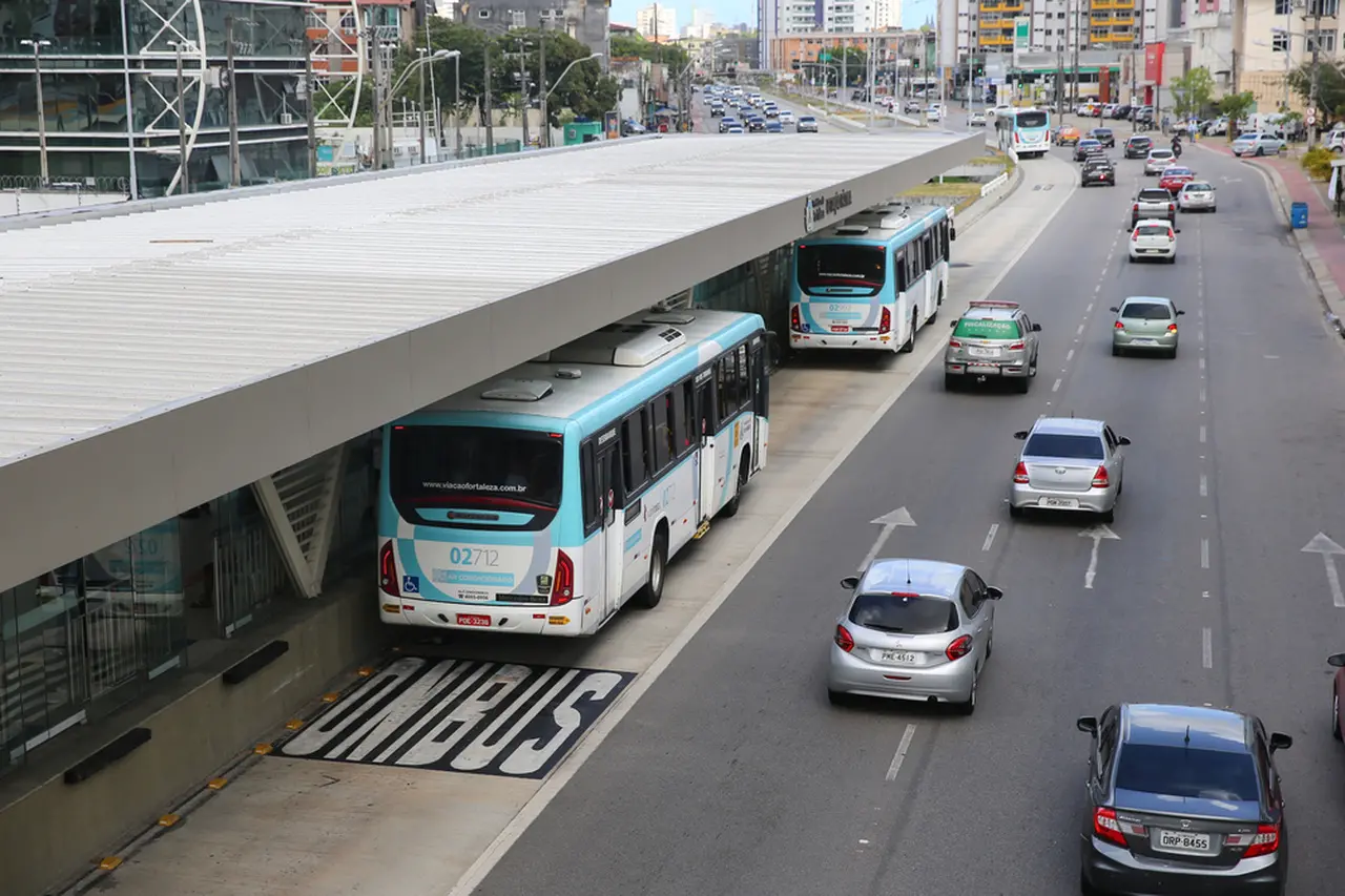 Imagem mostra BRT instalado na av. Aguananbi em Fortaleza, com vários ônibus brancos e azuis estacionados e carros passando ao redor na cidade durante o dia, com edifícios modernos ao fundo.