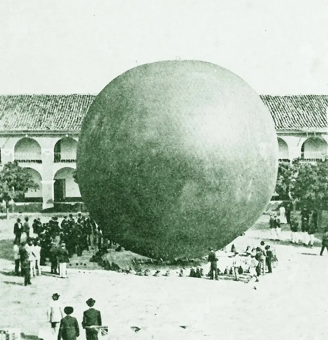 Fotografia antiga em tons de verde e branco mostrando um enorme balão esférico no pátio de um edifício colonial. Diversas pessoas em trajes de época observam a estrutura monumental ao redor.