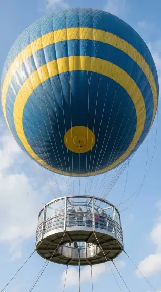 Vista inferior de um balão azul com listras amarelas. Uma cabine circular metálica e envidraçada, repleta de pessoas, está suspensa por cabos sob o balão contra um céu claro.