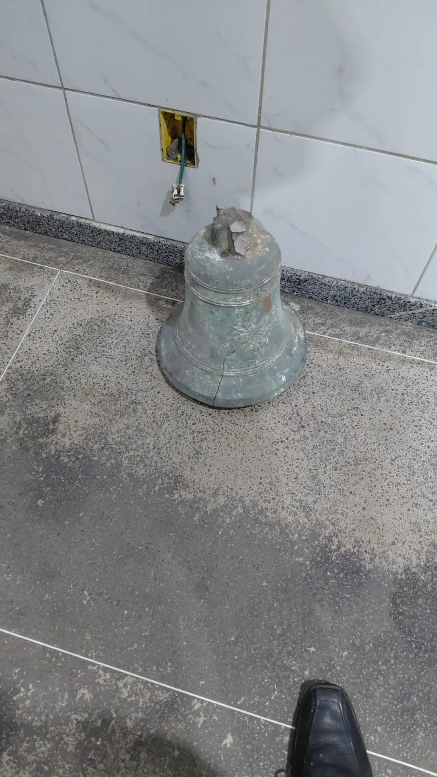 Um antigo sino de bronze com pátina esverdeada e uma rachadura visível repousa diretamente sobre um piso de granito cinza. Ao fundo, uma parede de azulejos brancos exibe uma tomada elétrica aberta e fios expostos.