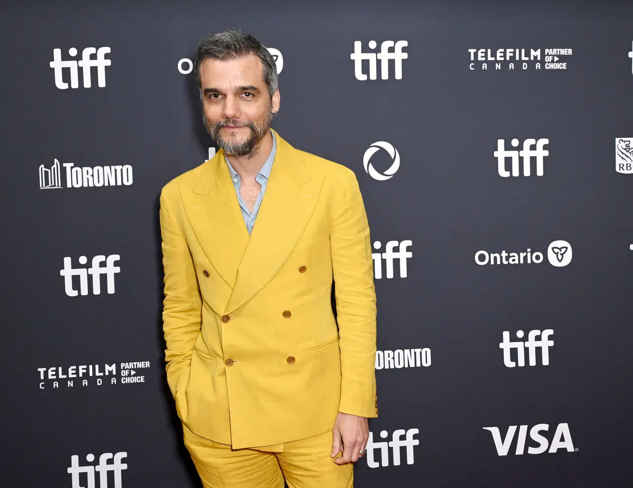 O ator Wagner Moura posa no tapete vermelho do Festival de Toronto usando um terno de linho amarelo vibrante sobre uma camisa social azul clara. Ele está posicionado à esquerda contra um fundo cinza repleto de logotipos do evento, mantendo uma expressão serena e as mãos nos bolsos.