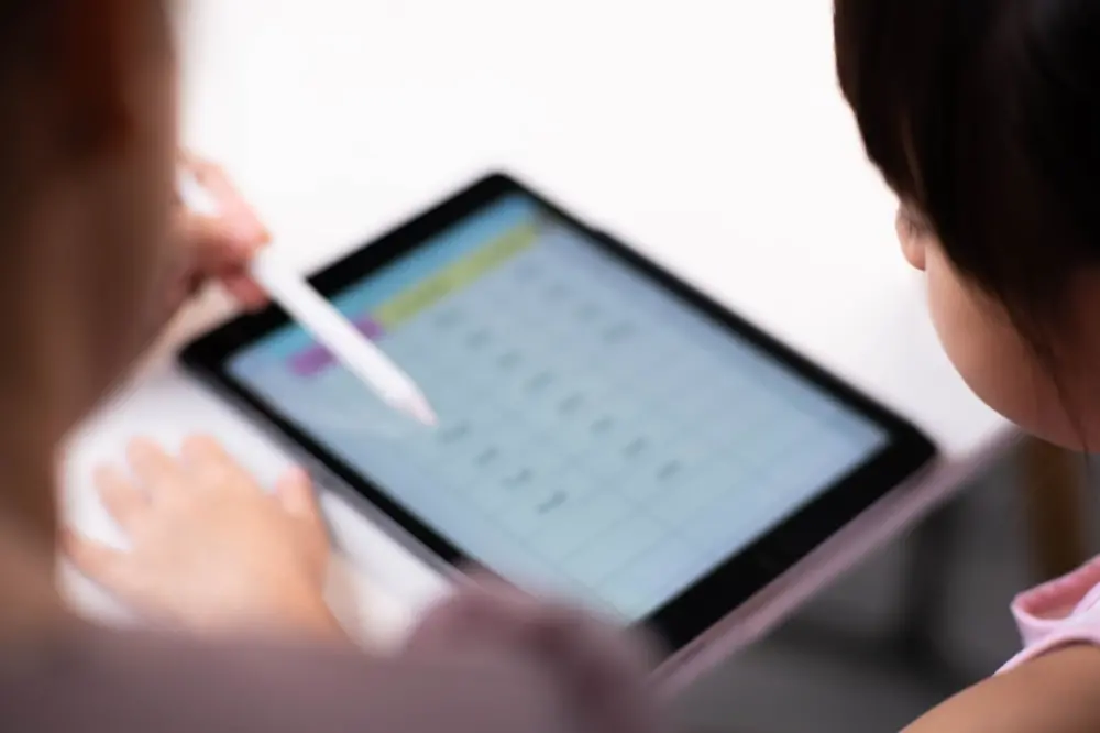 Na imagem, foto em ângulo superior e levemente desfocada mostrando duas crianças interagindo com um tablet sobre uma mesa branca. Uma das crianças segura uma caneta digital branca, apontando para a tela que exibe uma tabela ou atividade educativa. Apenas parte do rosto e das mãos das crianças é visível, com foco principal no uso da tecnologia no aprendizado.
