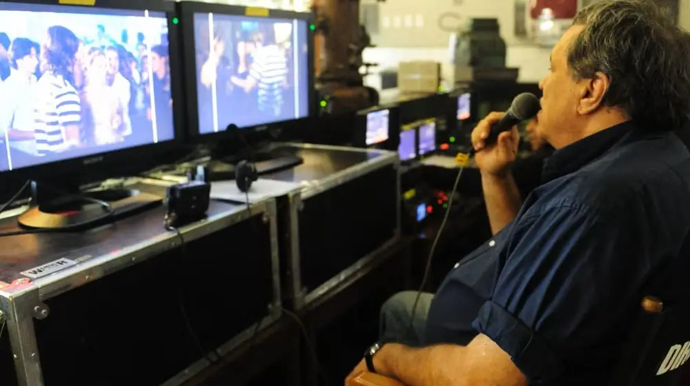 Na imagem, foto de perfil de Dennis Carvalho sentado em sua cadeira de diretor durante uma gravação. Ele segura um microfone próximo à boca, observando atentamente dois monitores de vídeo à sua frente. As telas exibem cenas de atores em um ambiente de festa. O diretor veste uma camisa azul escura de mangas curtas e está em uma área técnica com equipamentos de vídeo protegidos por cases pretos.