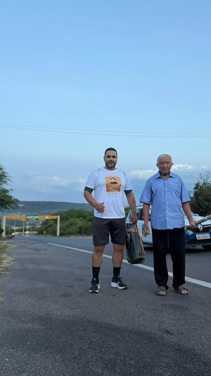 Na imagem, foto de corpo inteiro, em plano médio, de dois homens em pé no acostamento de uma rodovia durante o entardecer. À esquerda, um homem mais jovem, de barba curta, veste uma camiseta branca com uma estampa religiosa (a mesma da foto anterior), bermuda escura e tênis pretos; ele segura uma sacola plástica. À direita, um senhor idoso (Seu Osmundo) veste uma camisa social azul de mangas curtas com listras verticais finas, calça escura e sandálias; ele segura um objeto longo e cilíndrico, que parece ser um cajado ou suporte para bandeira.