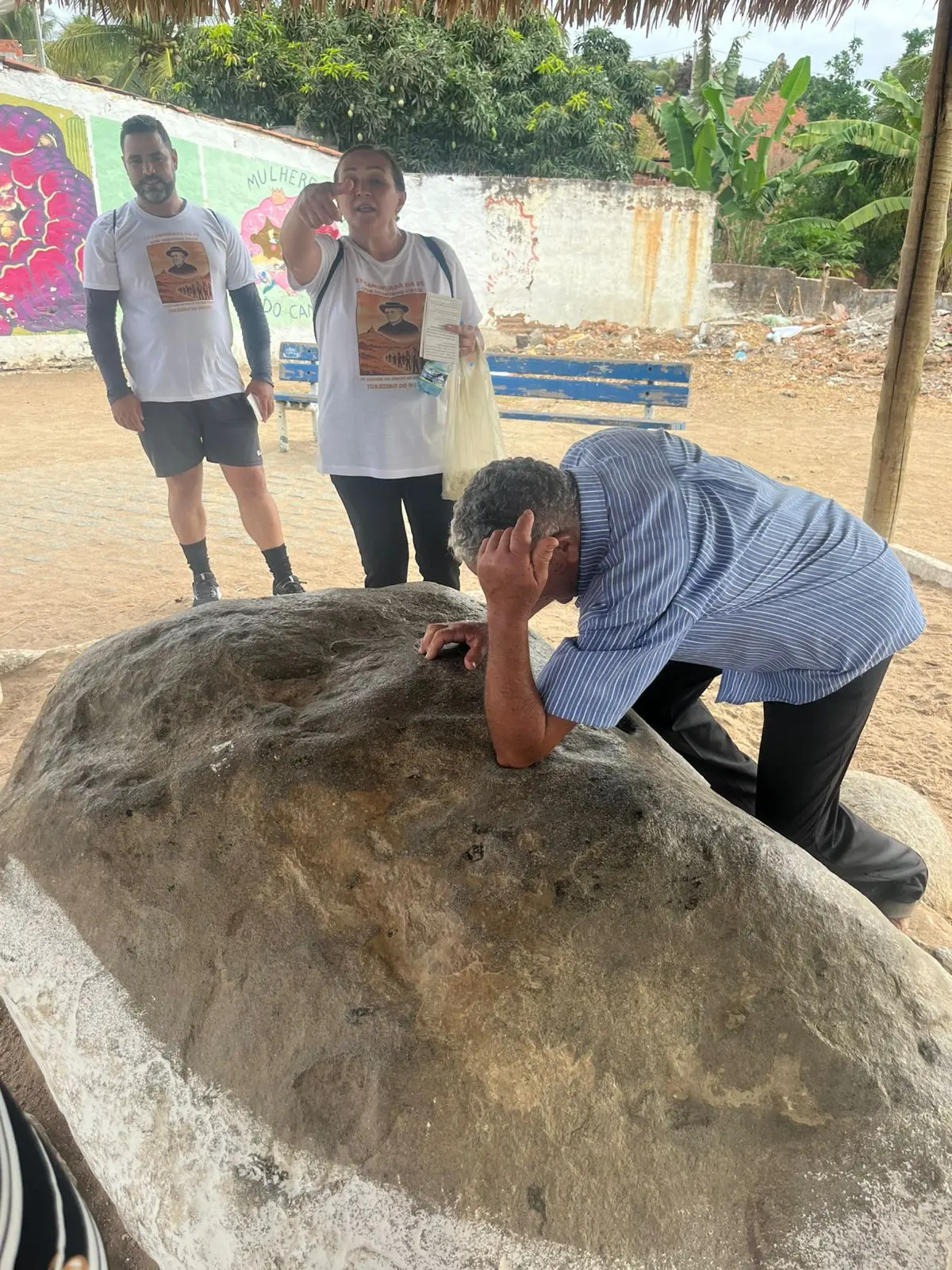 Na imagem, foto em plano médio tirada sob uma cobertura de palha. Em primeiro plano, Seu Osmundo está curvado sobre uma grande rocha arredondada, apoiando a cabeça e o braço sobre ela em um gesto de oração ou cansaço. Atrás dele, uma mulher e o mesmo homem jovem da foto anterior observam; a mulher aponta para algo fora do quadro enquanto segura folhetos. Ambos vestem as camisetas brancas com a estampa do Padre Cícero. Ao fundo, vê-se um muro com grafites coloridos e vegetação tropical, como bananeiras.