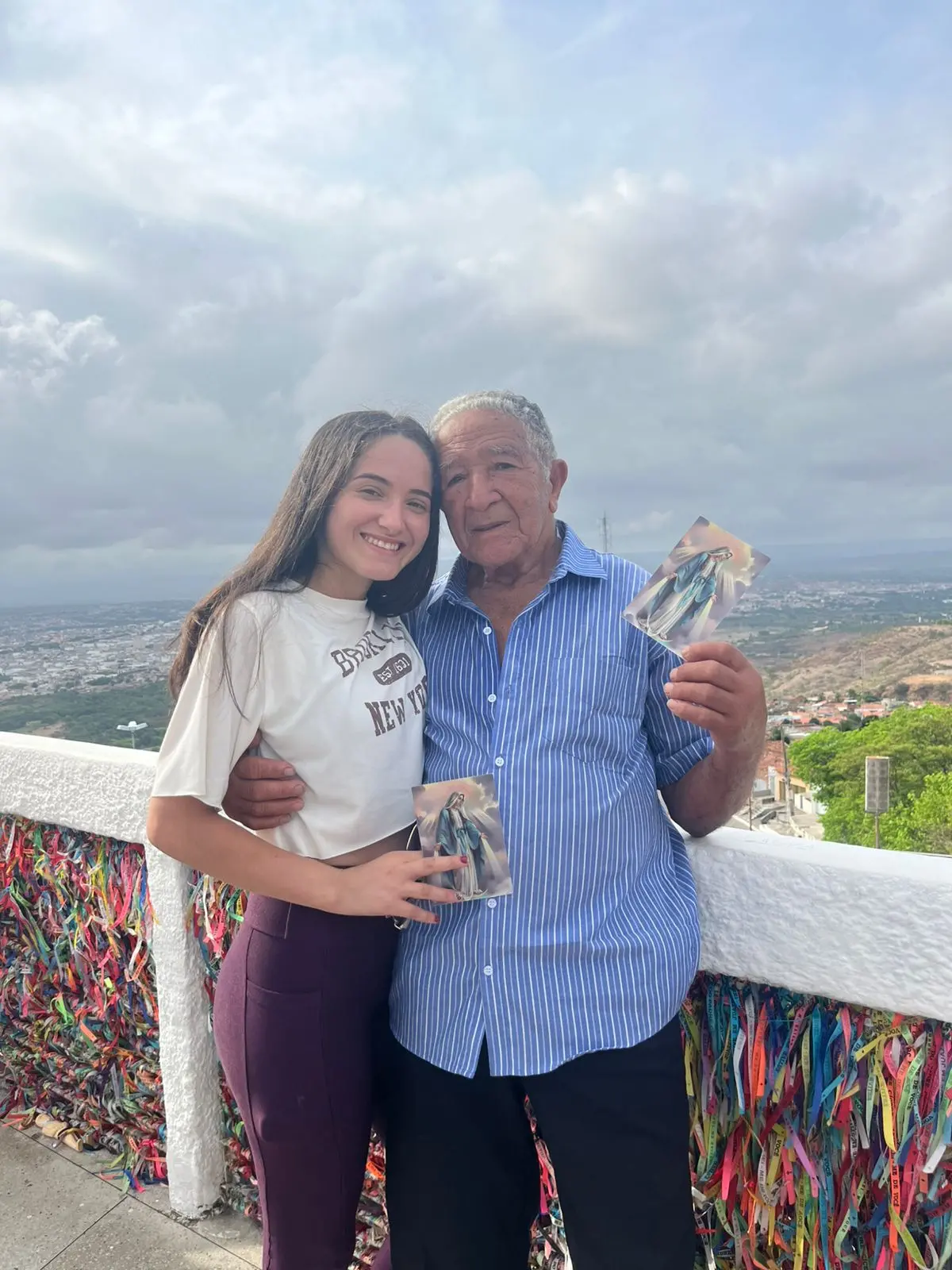 Na imagem, foto vertical em plano médio de Seu Osmundo e uma jovem sorridentes, abraçados em um mirante. Ele veste sua camisa social azul listrada e ela uma camiseta branca com calça vinho. Ambos seguram, cada um, um pequeno cartão com a imagem de Nossa Senhora das Graças. Eles estão encostados em uma mureta branca totalmente coberta por milhares de fitinhas coloridas de pedidos (fitas do Bonfim ou de promessas). Ao fundo, a vista panorâmica revela uma cidade vasta sob um céu nublado, indicando que chegaram ao topo de um destino de peregrinação.