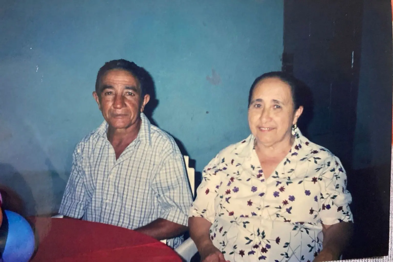 Osmundo José da Cruz e Maria Ivete Leite da Cruz. Casal idoso sorridente, ele de camisa xadrez e ela de blusa floral, sentados à mesa com parede azul ao fundo.
