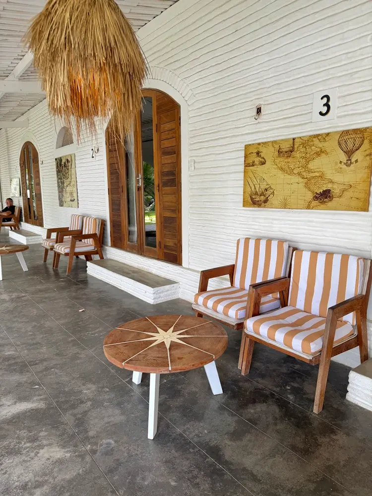 Na imagem, uma varanda externa de estilo rústico e sofisticado com paredes de tijolos brancos texturizados. O ambiente apresenta portas de madeira em arco e mobiliário de madeira que inclui poltronas com estofado listrado em branco e bege. À frente das poltronas, há uma mesa de centro redonda de madeira com uma marchetaria no topo representando uma rosa dos ventos.