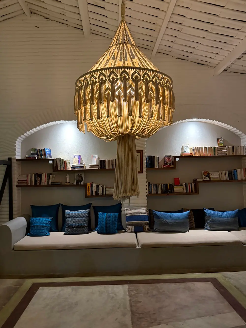 Na imagem, Uma varanda externa iluminada pela luz do dia, com paredes brancas de tijolos aparentes dispostos de forma horizontal. O espaço conta com pares de poltronas de madeira com estofamento listrado em bege e branco. À frente delas, destaca-se uma mesa de centro redonda de madeira clara com um desenho de rosa dos ventos em marchetaria no centro.