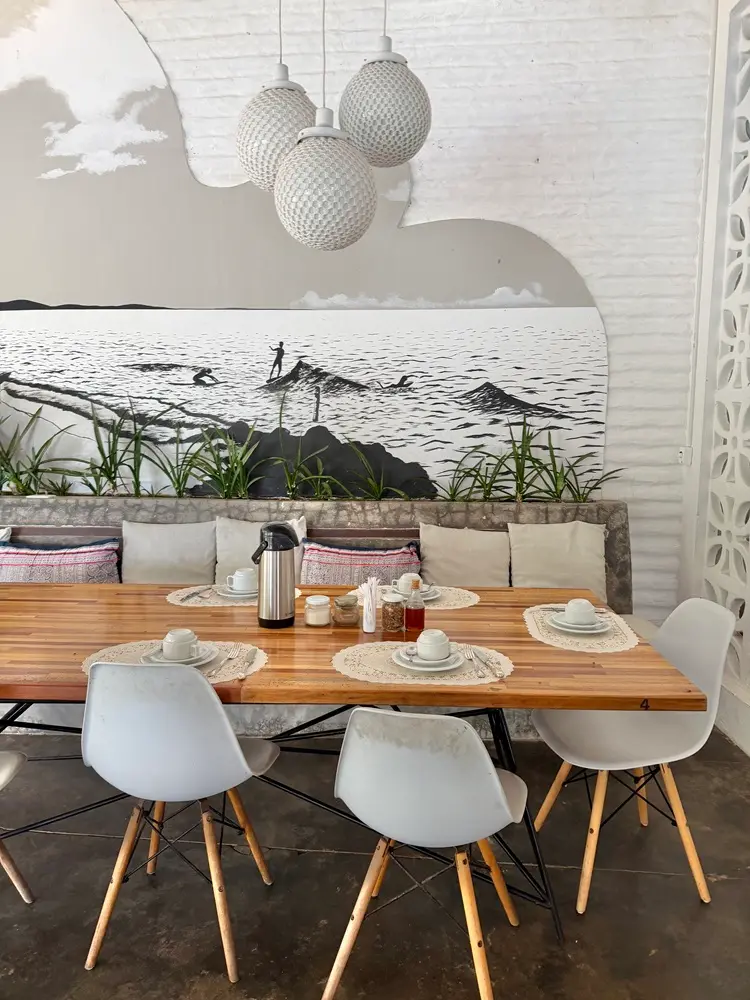 Na imagem, uma mesa de jantar comunitária de madeira clara, posta para o café da manhã, situada em um ambiente decorado com temática litorânea. Ao fundo, destaca-se um mural artístico que ocupa a parede de tijolos brancos, retratando uma cena de surfe em preto e branco com silhuetas de surfistas em ondas e rochas.