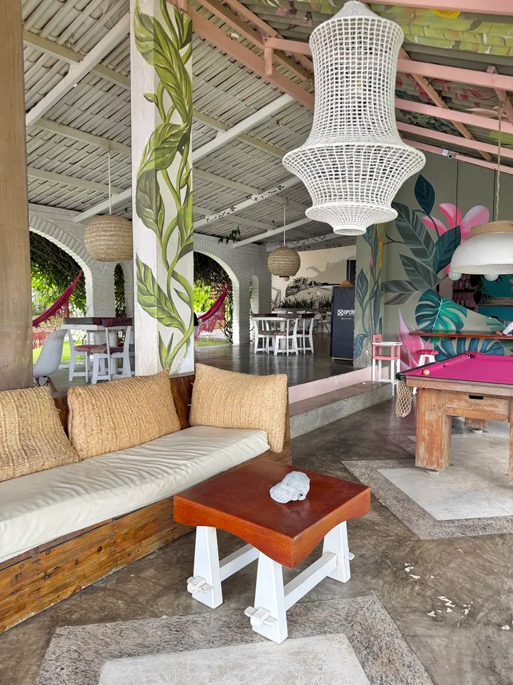 Na imagem, uma ampla área de lazer coberta e aberta nas laterais, com decoração tropical e rústica. O espaço combina móveis de madeira, iluminação artesanal e paredes decoradas com pinturas botânicas coloridas.