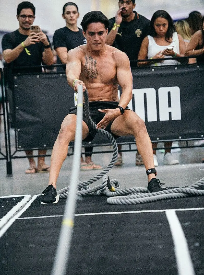 Pessoa sentada no chão durante uma prova funcional, segurando uma corda grossa com ambas as mãos e puxando-a em direção ao corpo. A pessoa está sem camisa, usando shorts escuros e tênis, com um grande desenho tatuado no peito. Ao fundo, há uma barreira de proteção e várias outras pessoas em pé observando ou registrando a cena com celulares. O ambiente parece ser um evento esportivo indoor.