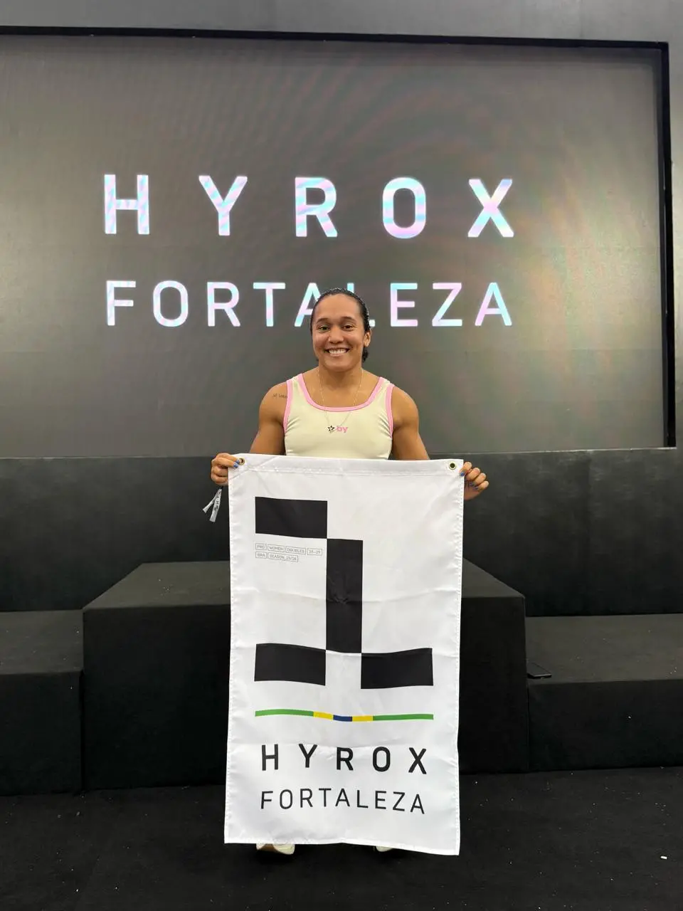 Pessoa em pé sobre um palco preto segurando uma bandeira branca com o logotipo da Hyrox Fortaleza, composta por formas geométricas pretas e uma linha colorida na base. Ao fundo, há um telão grande exibindo as palavras 'HYROX FORTALEZA' em letras brancas. A pessoa veste uma camiseta clara e está posicionada no centro da cena, com o palco e o telão ocupando a maior parte do ambiente.