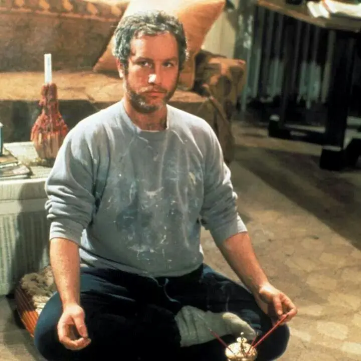 O ator Richard Dreyfuss aparece sentado de pernas cruzadas no chão em cena de 'A Garota do Adeus', vestindo um moletom cinza sujo de tinta e calças escuras em um ambiente doméstico. Ele encara a câmera com uma expressão séria, posicionado atrás de um pequeno queimador de incenso aceso que libera uma leve fumaça.