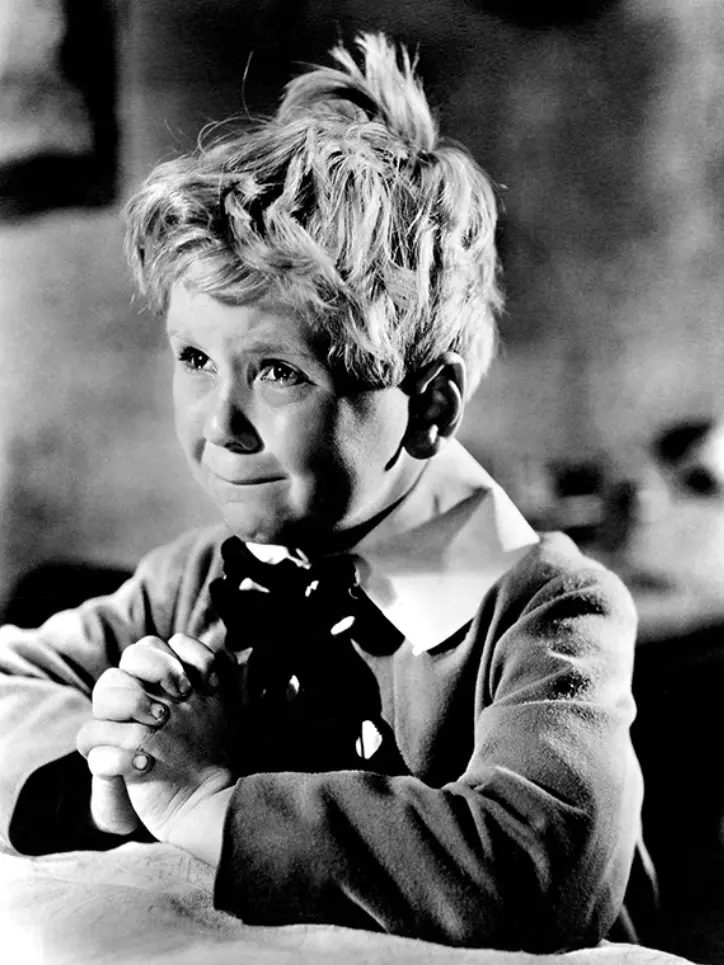 O ator mirim Jackie Cooper aparece em uma cena em preto e branco do filme 'Skippy', com as mãos postas em oração e uma expressão de profunda angústia e choro contido. Ele usa um casaco escuro sobre uma gola branca larga e um laço, destacando seu cabelo loiro bagunçado em uma composição emocionante que foca em seu rosto expressivo.