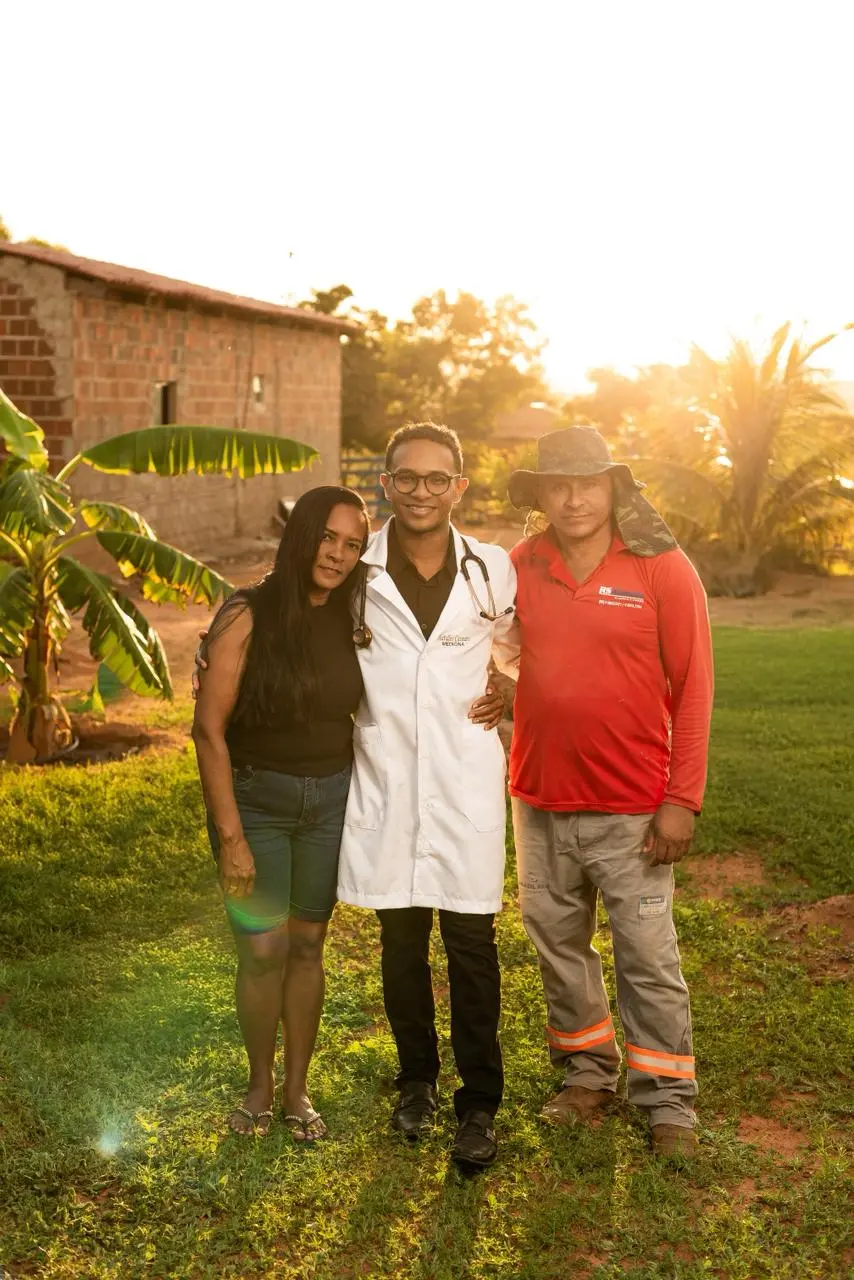 Na imagem, uma fotografia vertical ao ar livre, iluminada pela luz quente e dourada do pôr do sol, capturando três pessoas abraçadas em um terreno gramado. Ao centro: O mesmo jovem médico da imagem anterior, Nattalos Casseano. Ele sorri para a câmera, usando seu jaleco branco, camisa preta, calça social escura e estetoscópio no pescoço. Ele está com os braços ao redor dos ombros das duas pessoas ao seu lado. À esquerda: Uma mulher negra de cabelos longos e pretos, vestindo uma regata preta, bermuda jeans e sandálias rasteiras. Ela sorri levemente e abraça o jovem pela cintura. À direita: Um homem de pele clara, vestindo uma camisa de manga longa vermelha com um logotipo no peito, calças de trabalho cinza com faixas refletoras e um chapéu de sol camuflado com proteção de nuca. Ele sorri de forma calorosa. Ao fundo: O cenário é rural, com uma casa de tijolos aparentes à esquerda, uma bananeira e outras árvores ao fundo. O sol baixo cria um efeito de brilho suave e sombras longas na grama, transmitindo uma sensação de orgulho e união familiar.