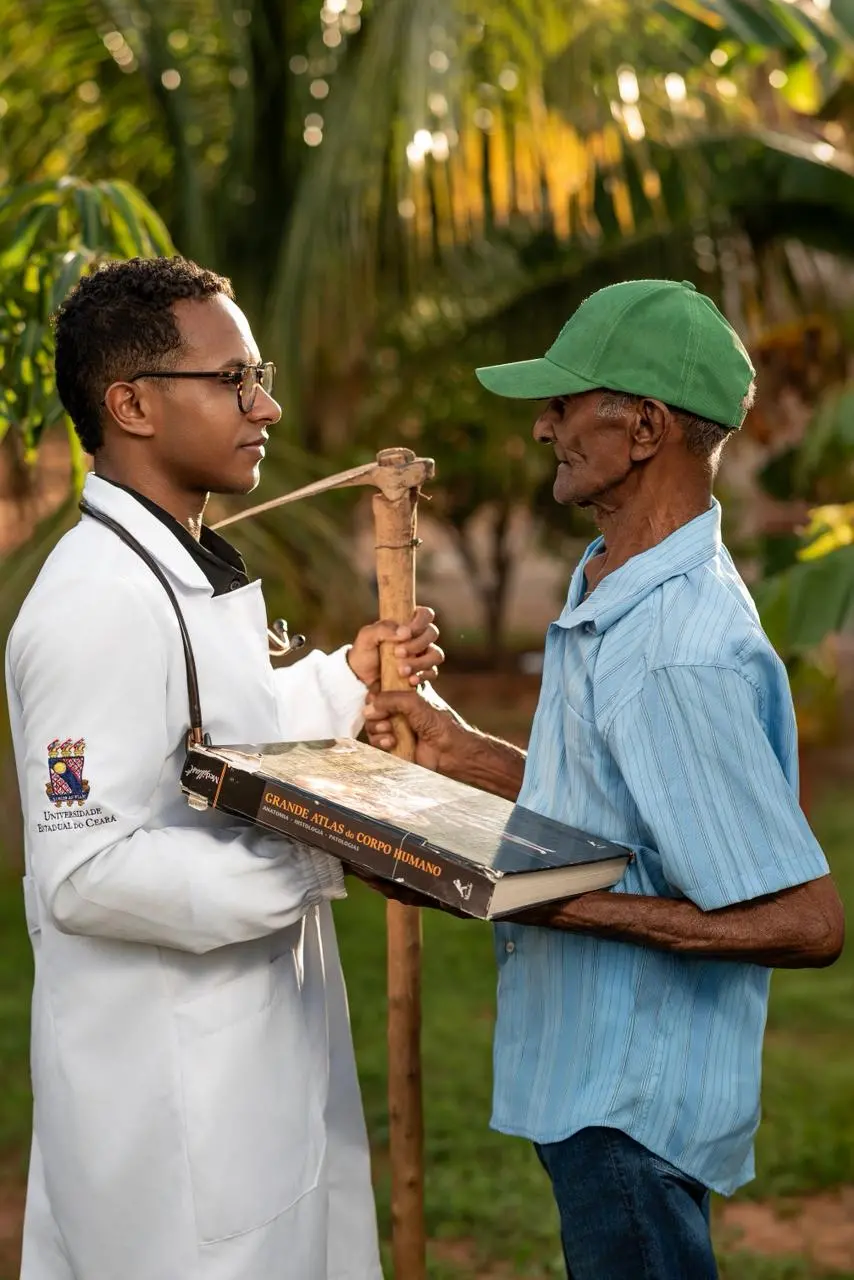 Na imagem, uma fotografia de plano médio, ao ar livre, mostrando três pessoas em pé sobre um gramado, iluminadas pela luz suave e dourada do final de tarde. No centro, o jovem médico negro, Nattalos Casseano, sorri e abraça pelos ombros as duas pessoas ao seu lado. Ele veste seu jaleco branco com o logotipo da 