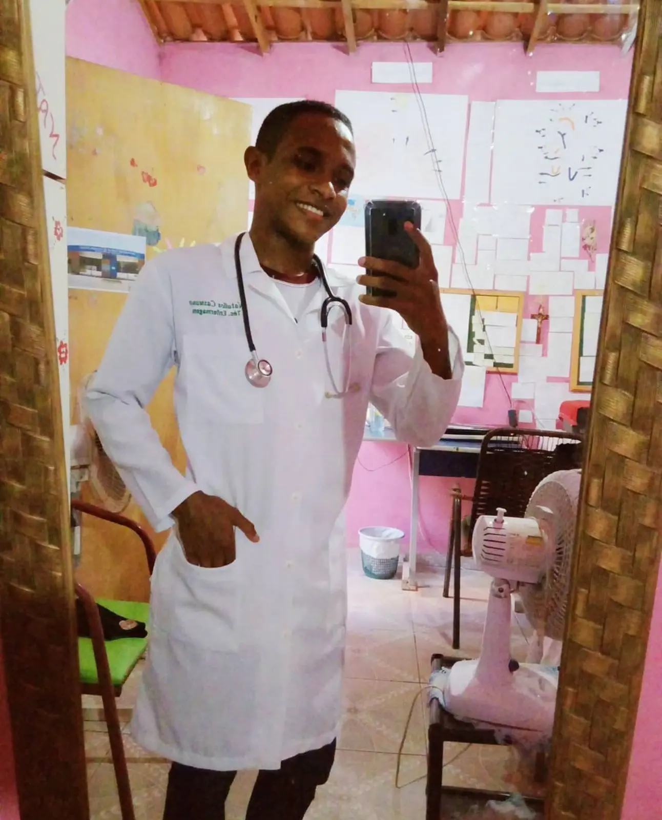 Na imagem, uma selfie tirada em frente a um espelho com moldura trançada, em um ambiente interno simples com paredes cor-de-rosa. O jovem Nattalos aparece sorrindo, vestindo um jaleco branco com seu nome bordado em verde e um estetoscópio no pescoço. Ele está com uma mão no bolso do jaleco e segura o celular com a outra. Ao fundo, o quarto possui materiais de estudo colados na parede, um ventilador de mesa e uma cadeira verde, evidenciando um ambiente de dedicação aos estudos.