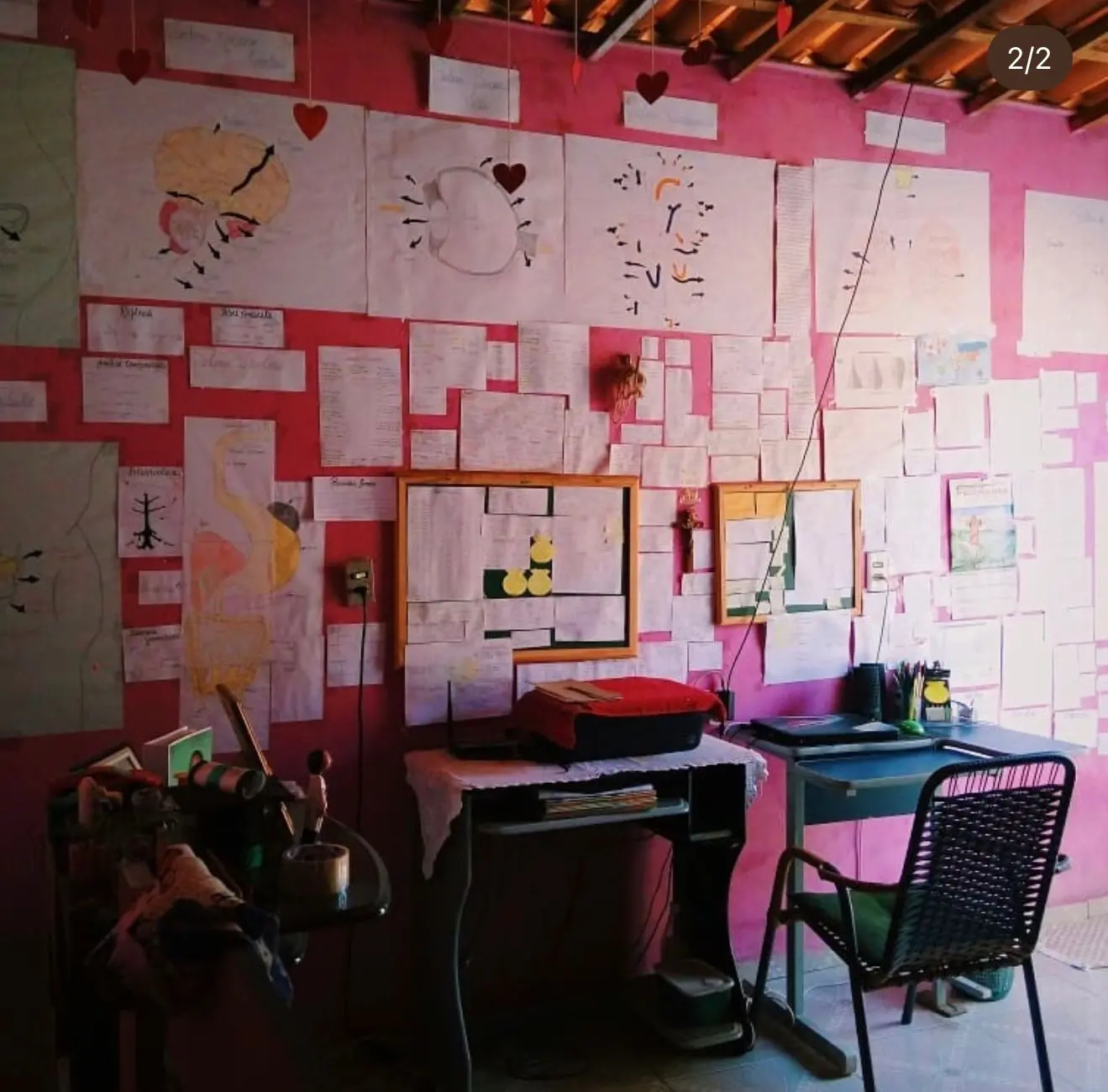 Na imagem, uma foto em plano aberto de um espaço de estudos improvisado dentro de um quarto com paredes rosa e teto de telha aparente. A parede principal está quase inteiramente coberta por cartazes feitos à mão com desenhos detalhados de órgãos humanos (coração, estômago, cérebro) e muitas folhas de papel com anotações de estudo. À frente da parede, há uma mesa simples com uma impressora e um notebook, uma cadeira de fios verde e pequenos enfeites de coração pendurados no teto. É um retrato visual de dedicação extrema e foco.