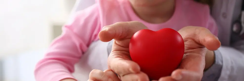 Na imagem, foto em close-foco aproximado mostrando as mãos de um adulto e de uma criança segurando juntas um coração vermelho de brinquedo. Ao fundo, de forma levemente desfocada, a criança veste uma blusa rosa e o adulto usa um jaleco branco com um estetoscópio pendurado no pescoço, sugerindo um ambiente de cuidado médico pediátrico.