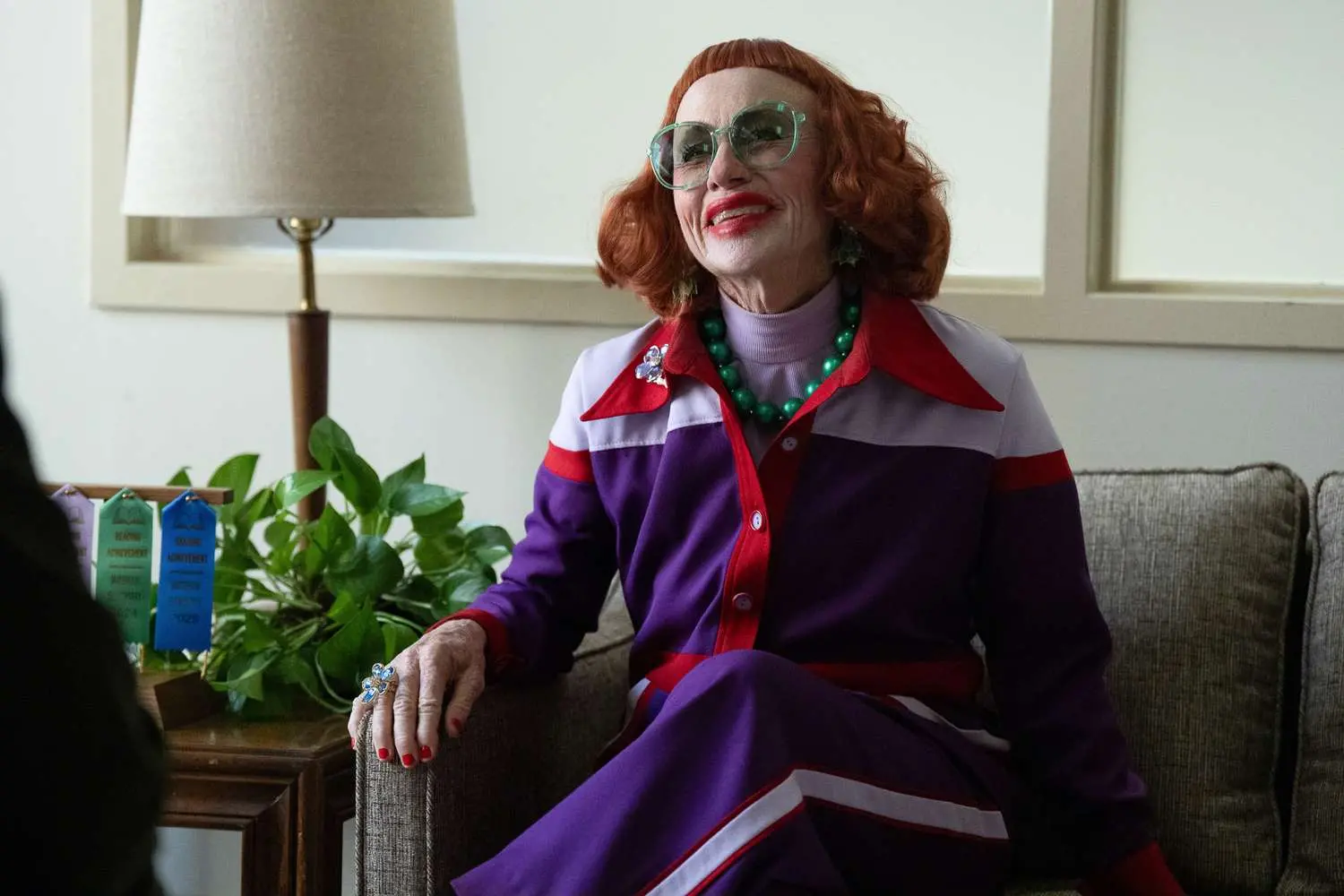 Amy Madigan, caracterizada como a personagem Aunt Gladys, sorri sentada em um sofá usando um conjunto roxo vibrante com detalhes em vermelho e branco, óculos de armação verde e um colar de contas grandes. Seu visual excêntrico é completado por cabelos ruivos ondulados e uma maquiagem marcante, em uma composição de cores vivas que contrasta com o ambiente doméstico ao fundo.