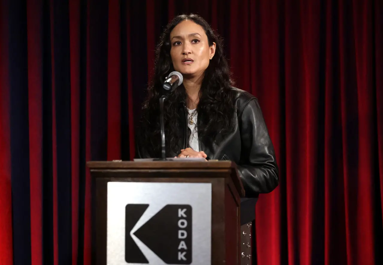 A diretora de fotografia Autumn Durald Arkapaw fala ao microfone atrás de um púlpito com a logomarca da Kodak, vestindo uma jaqueta de couro preta e com seus longos cabelos escuros soltos. O fundo é composto por cortinas vermelhas profundas que criam um cenário solene e focado durante o evento.